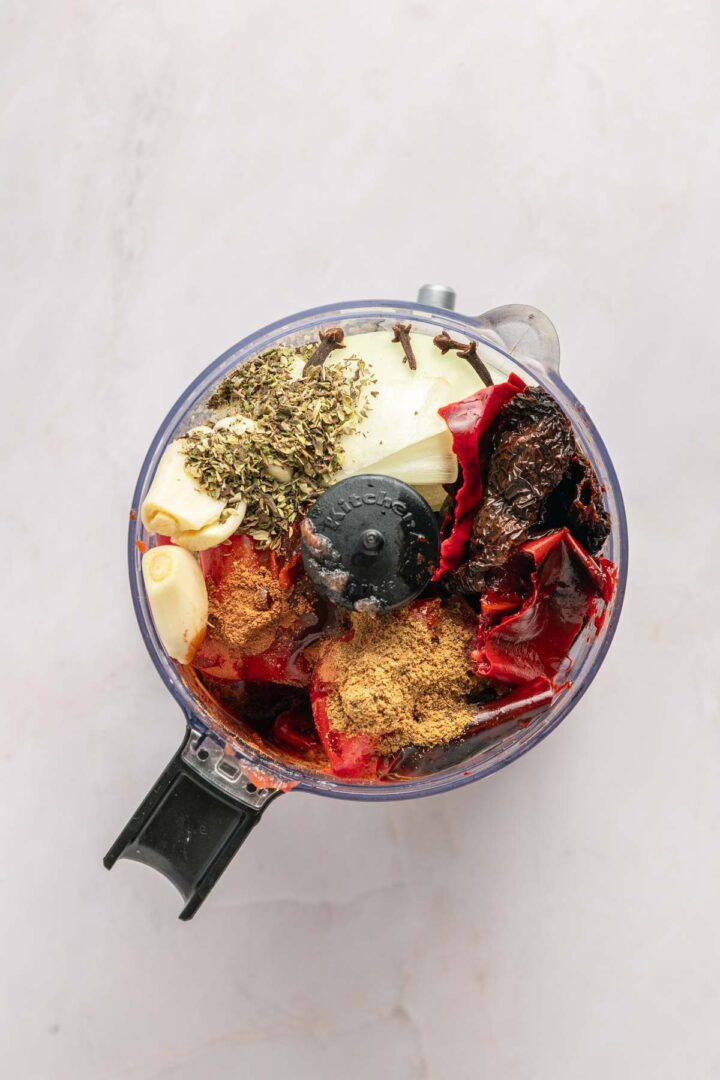 Overhead view of a food processor filled with peppers, garlic, onion, dried herbs, and spices&mdash;perfect for adding bold flavor to Birria Tacos or slow cooker taco recipes.