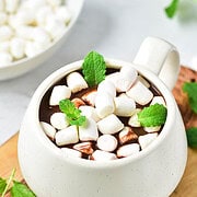 A white mug of peppermint hot chocolate topped with mini marshmallows and mint leaves, served on a wooden board.