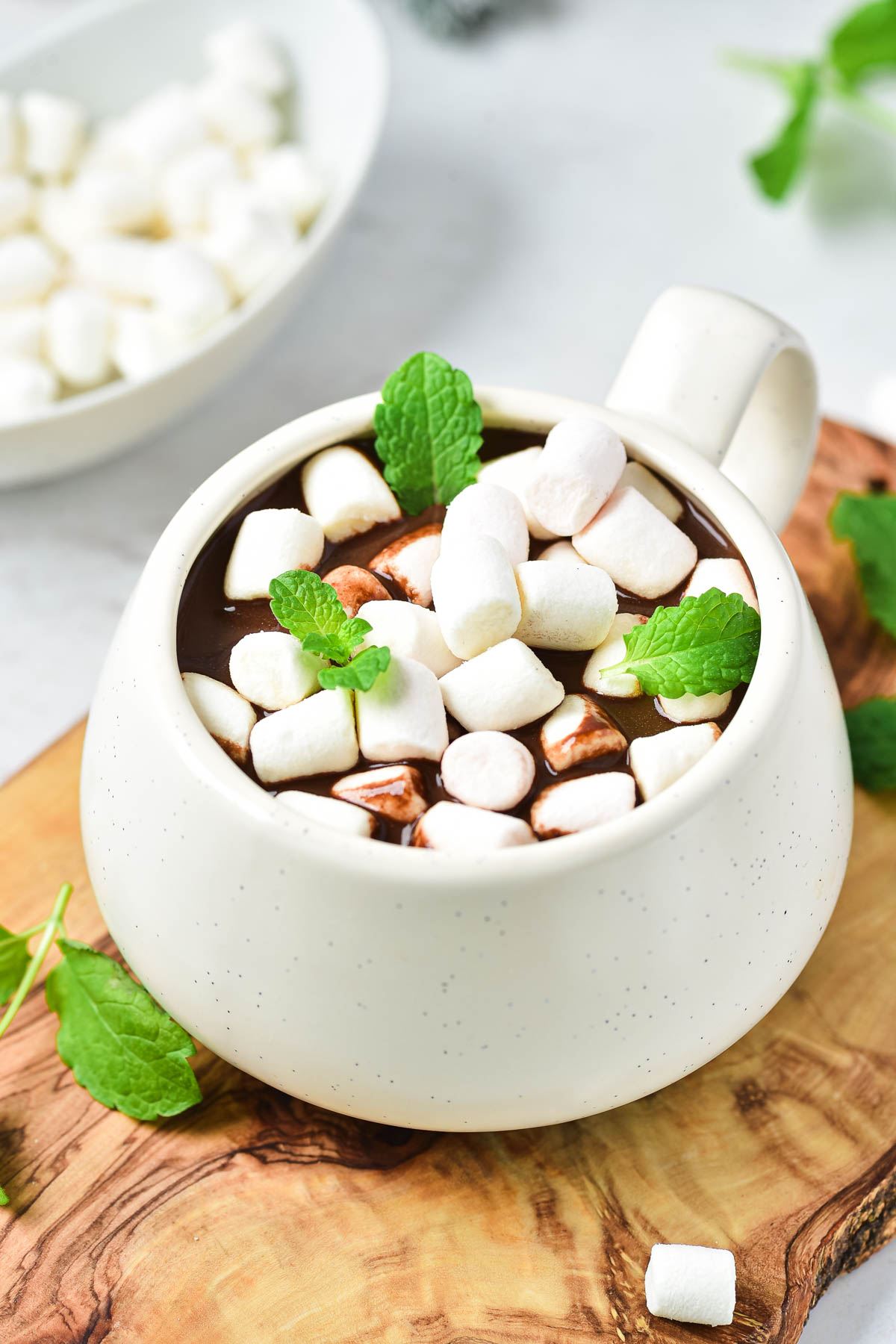 A white mug of peppermint hot chocolate topped with mini marshmallows and mint leaves, served on a wooden board.