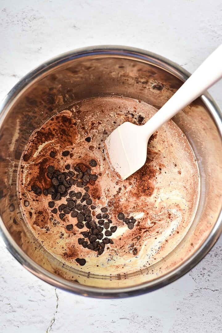 A mixing bowl with cream, cocoa powder, chocolate chips, and a white spatula on a light surface&mdash;perfect ingredients for making Peppermint Hot Chocolate.