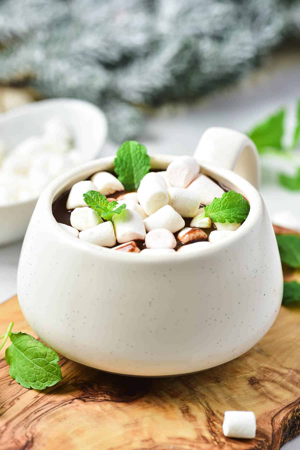 A mug of Peppermint Hot Chocolate topped with mini marshmallows and mint leaves sits on a wooden board.