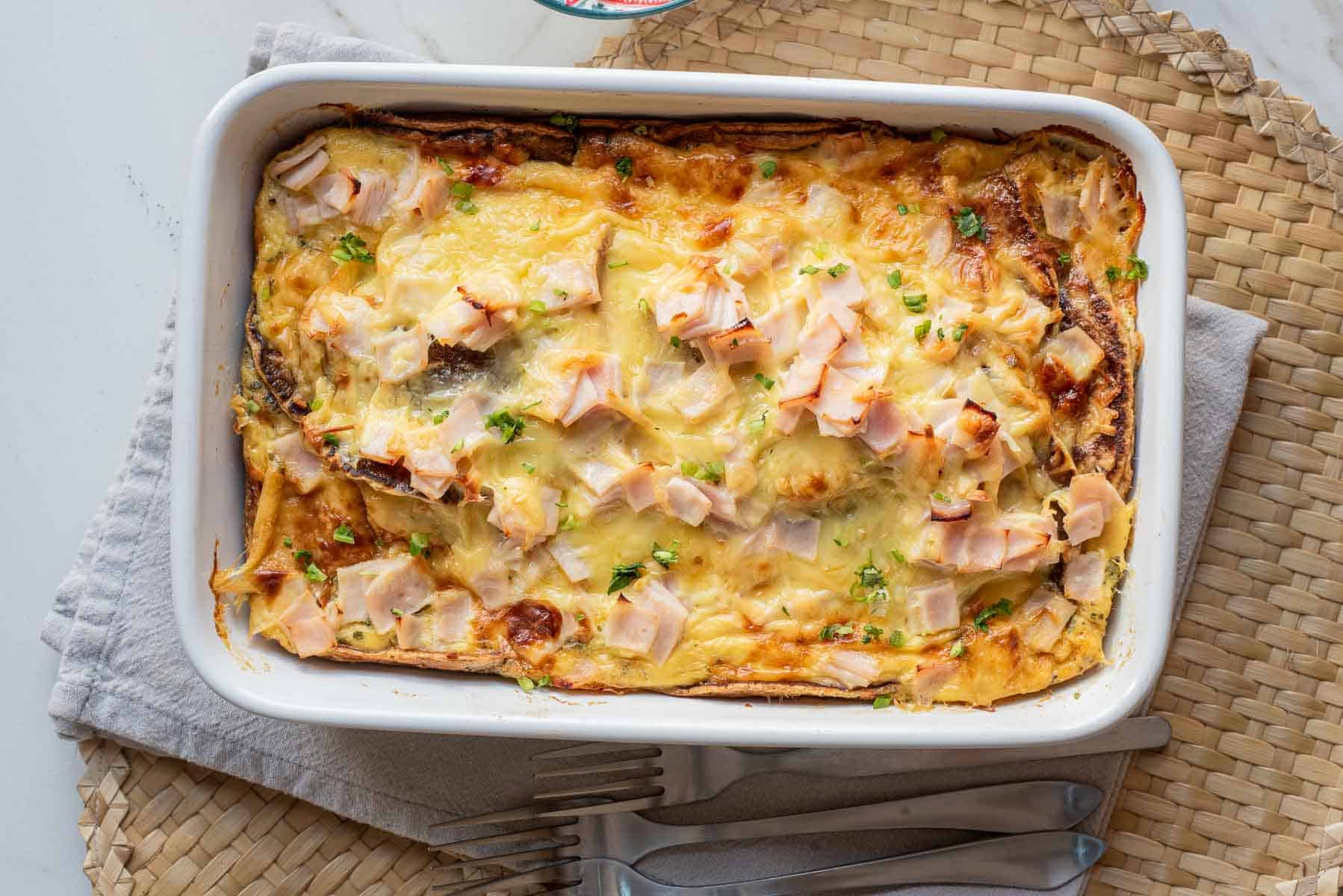 Baked breakfast casserole with melted cheese and chopped ham in a white dish on a woven placemat.