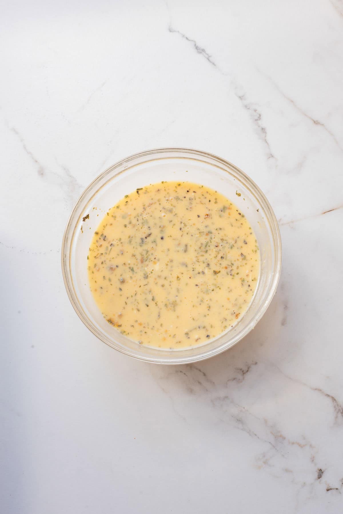A glass bowl filled with seasoned beaten eggs, perfect for a pancake recipe, sits on a white marble surface.