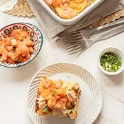 A slice of breakfast casserole topped with diced tomatoes sits on a plate, with more casserole and tasty toppings nearby—perfect for a hearty morning recipe.