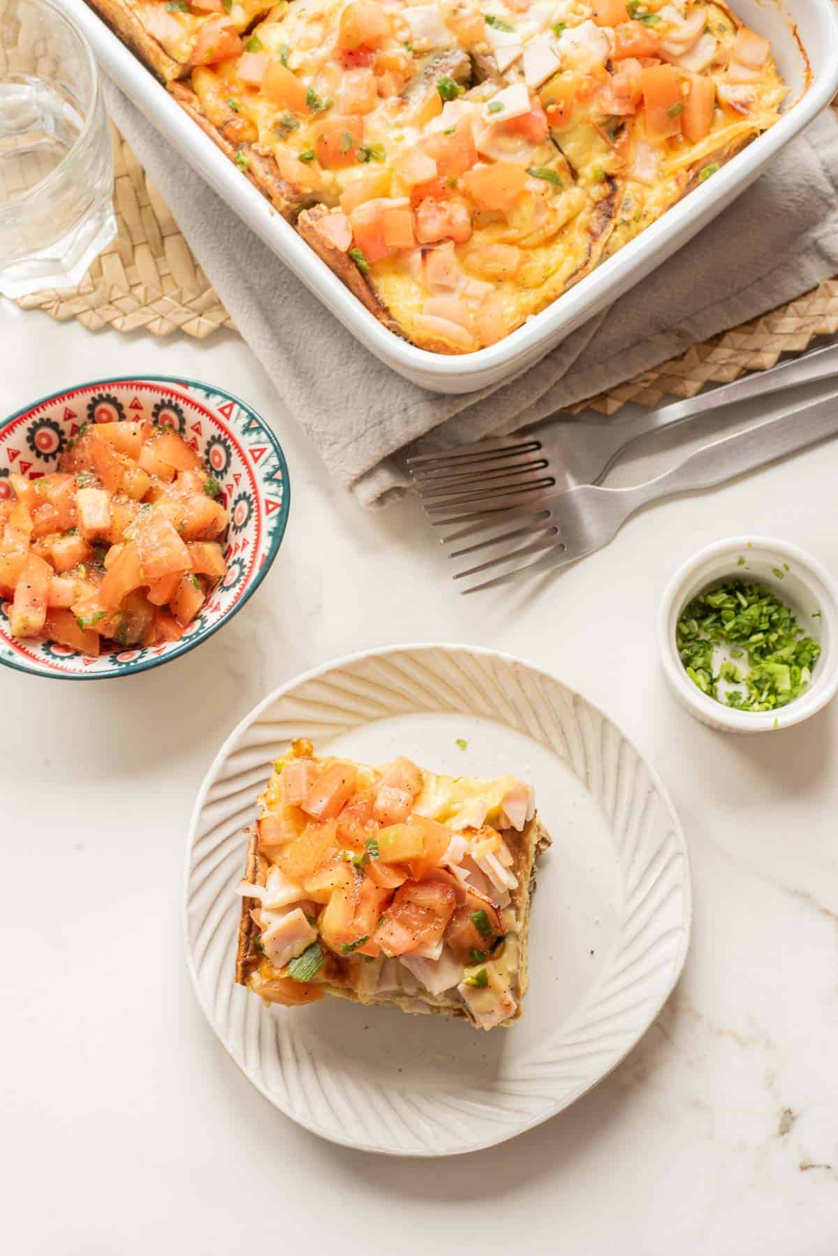 A slice of breakfast casserole topped with diced tomatoes sits on a plate, with more casserole and tasty toppings nearby&mdash;perfect for a hearty morning recipe.