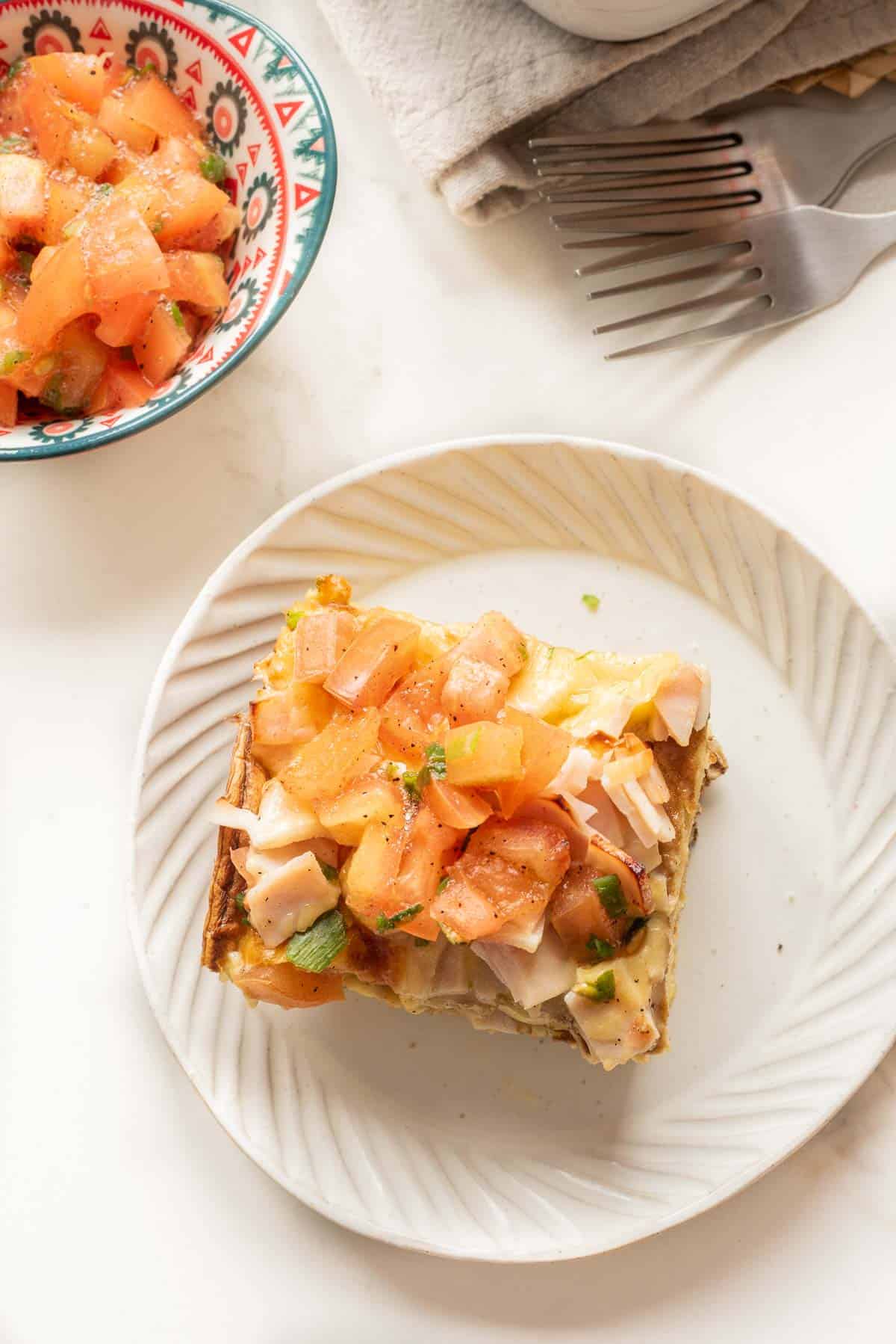 A white plate with a slice of savory Pancake Breakfast Casserole topped with diced tomatoes and herbs, beside a bowl of tomatoes.