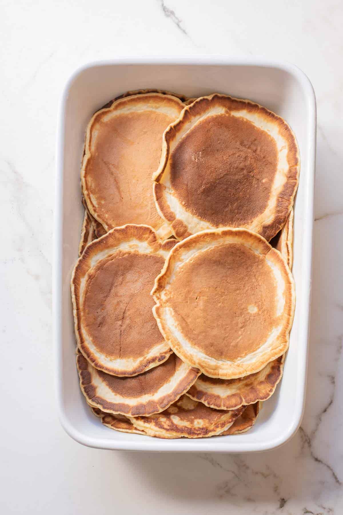 A white dish filled with a stack of golden brown pancakes on a marble surface, perfect inspiration for your next Pancake Breakfast Casserole.