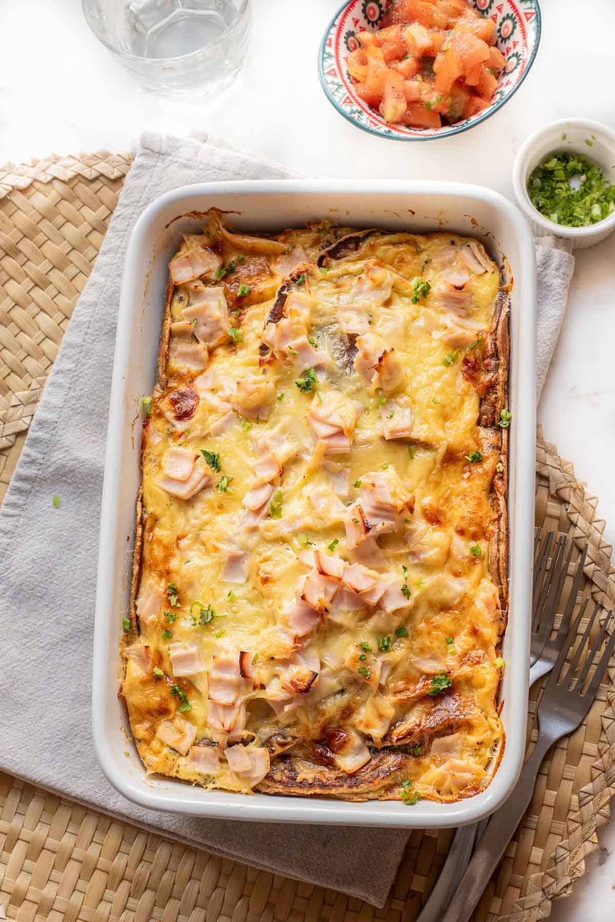 Breakfast casserole topped with melted cheese and diced ham, served on a woven placemat with salsa on the side&mdash;a delicious recipe that&rsquo;s perfect for starting your day.