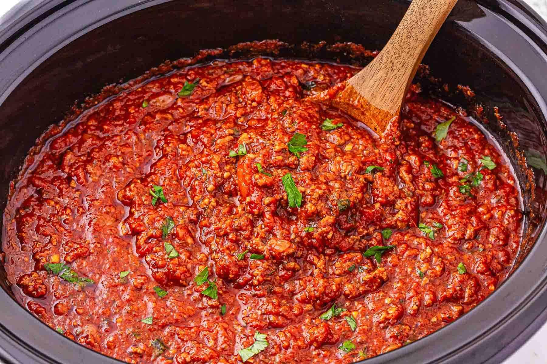 Thick red spaghetti Bolognese sauce with herbs is being stirred in a crockpot with a wooden spoon.