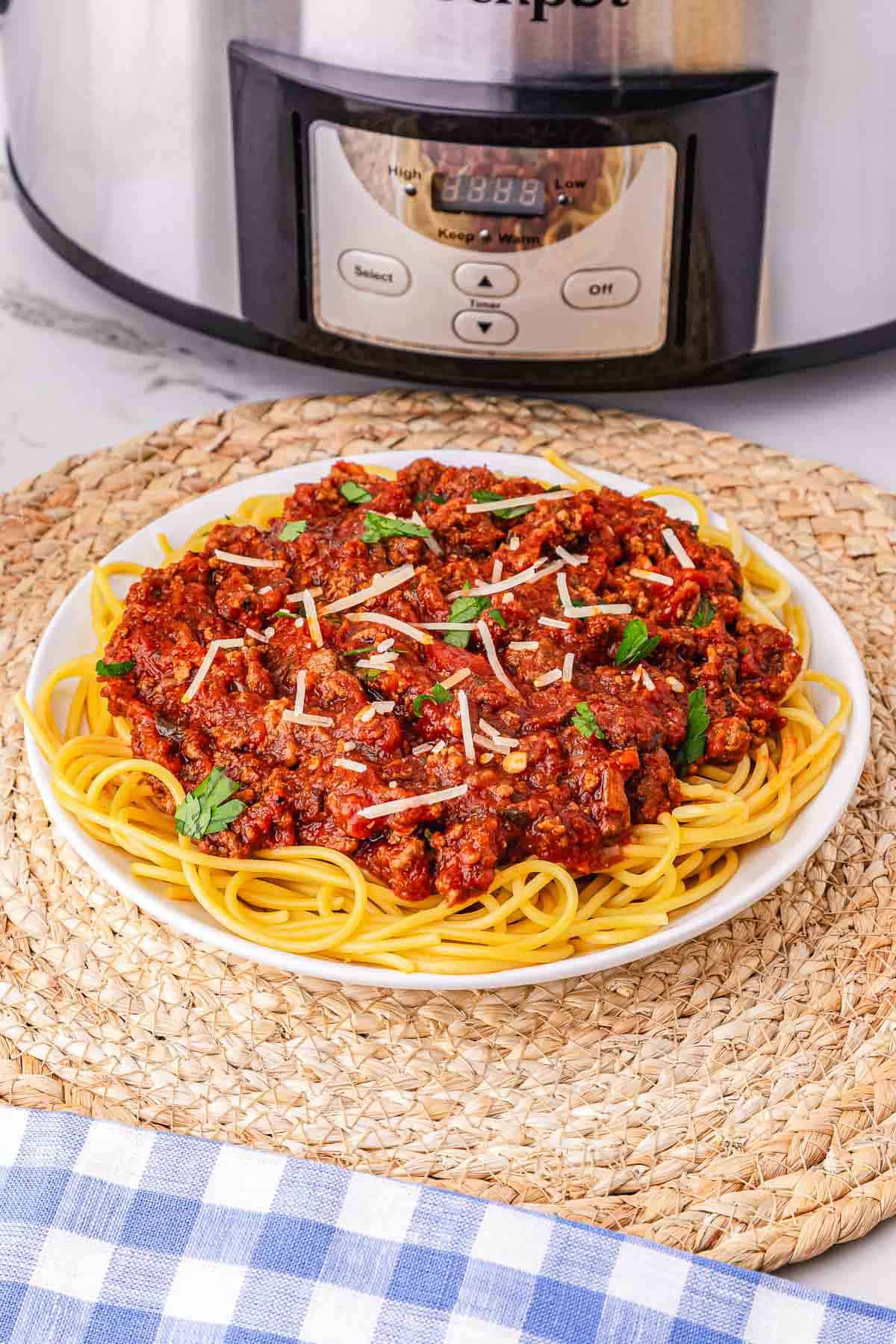 A plate of spaghetti Bolognese with rich meat sauce and cheese sits in front of a Crockpot on a woven placemat.