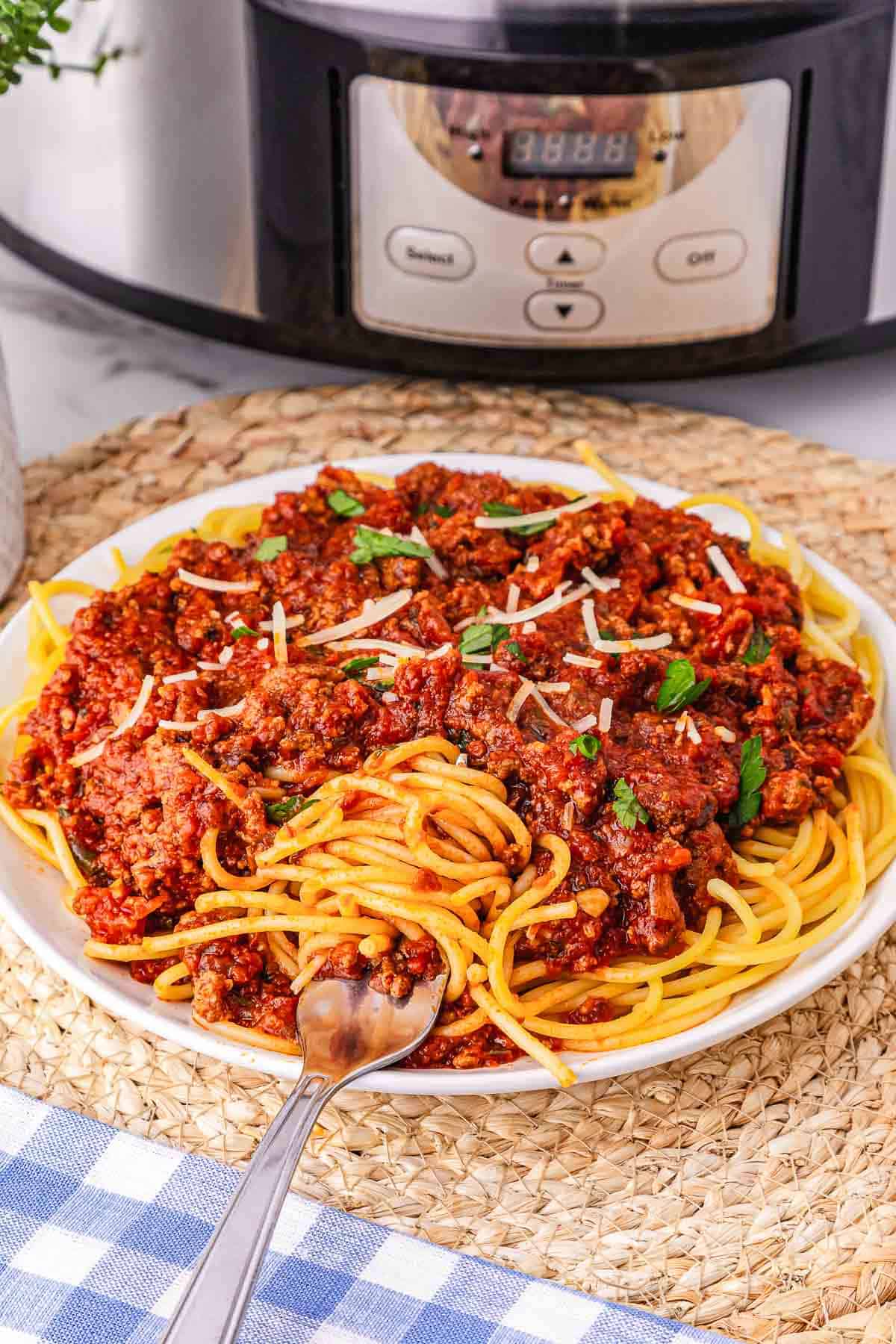 A plate of Spaghetti Bolognese with savory meat sauce, garnished with cheese and herbs, is served in front of a Crockpot.