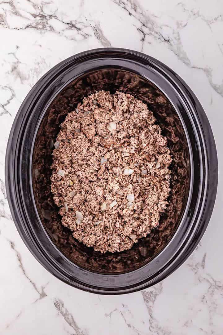 Ground beef and chopped onions simmer in a black Crockpot on a marble countertop, perfect for creating a flavorful meat sauce.