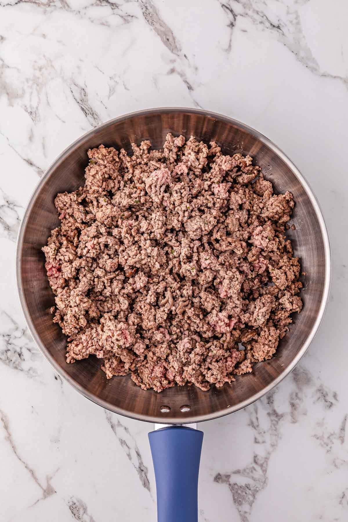 Ground beef cooking in a stainless steel skillet with a blue handle on a marble countertop, perfect for starting a rich Meat Sauce or Spaghetti Bolognese.