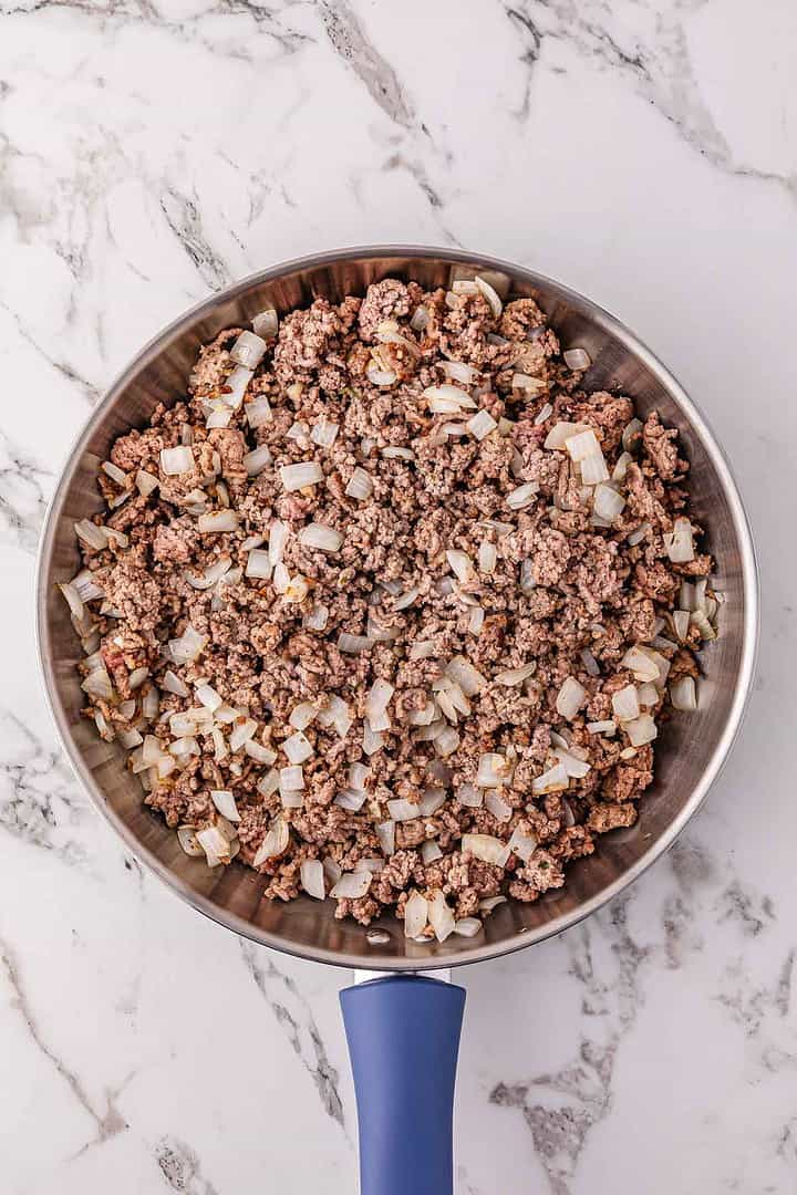 A skillet with cooked ground beef and chopped onions on a marble countertop, perfect for starting a hearty spaghetti bolognese.
