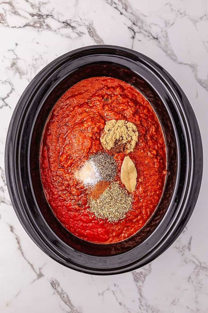 Rich spaghetti Bolognese meat sauce simmers in a Crockpot, topped with various spices and a bay leaf, on a marble countertop.
