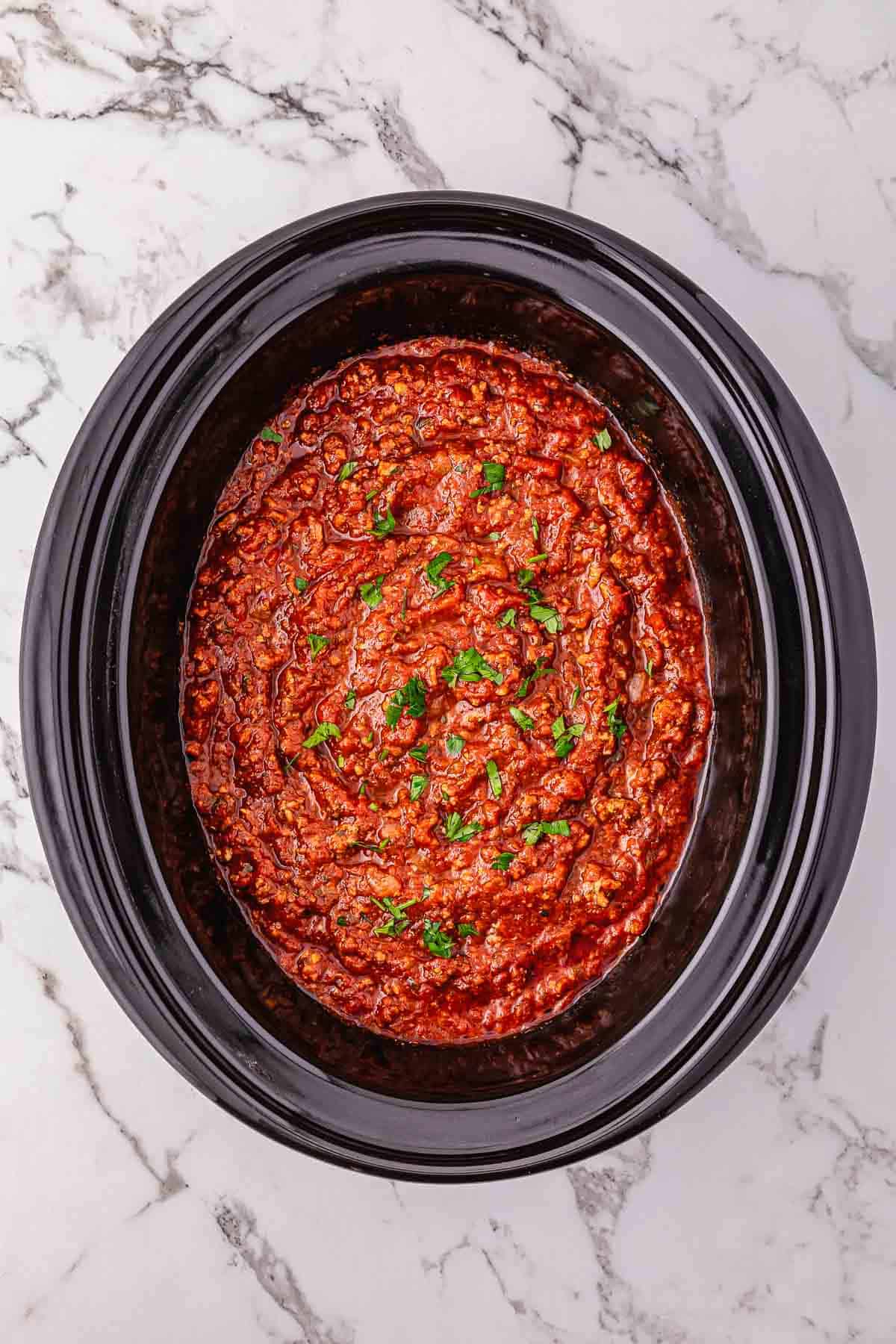 Hearty meat sauce with herbs simmers in a black crockpot, viewed from above on a white marble surface.