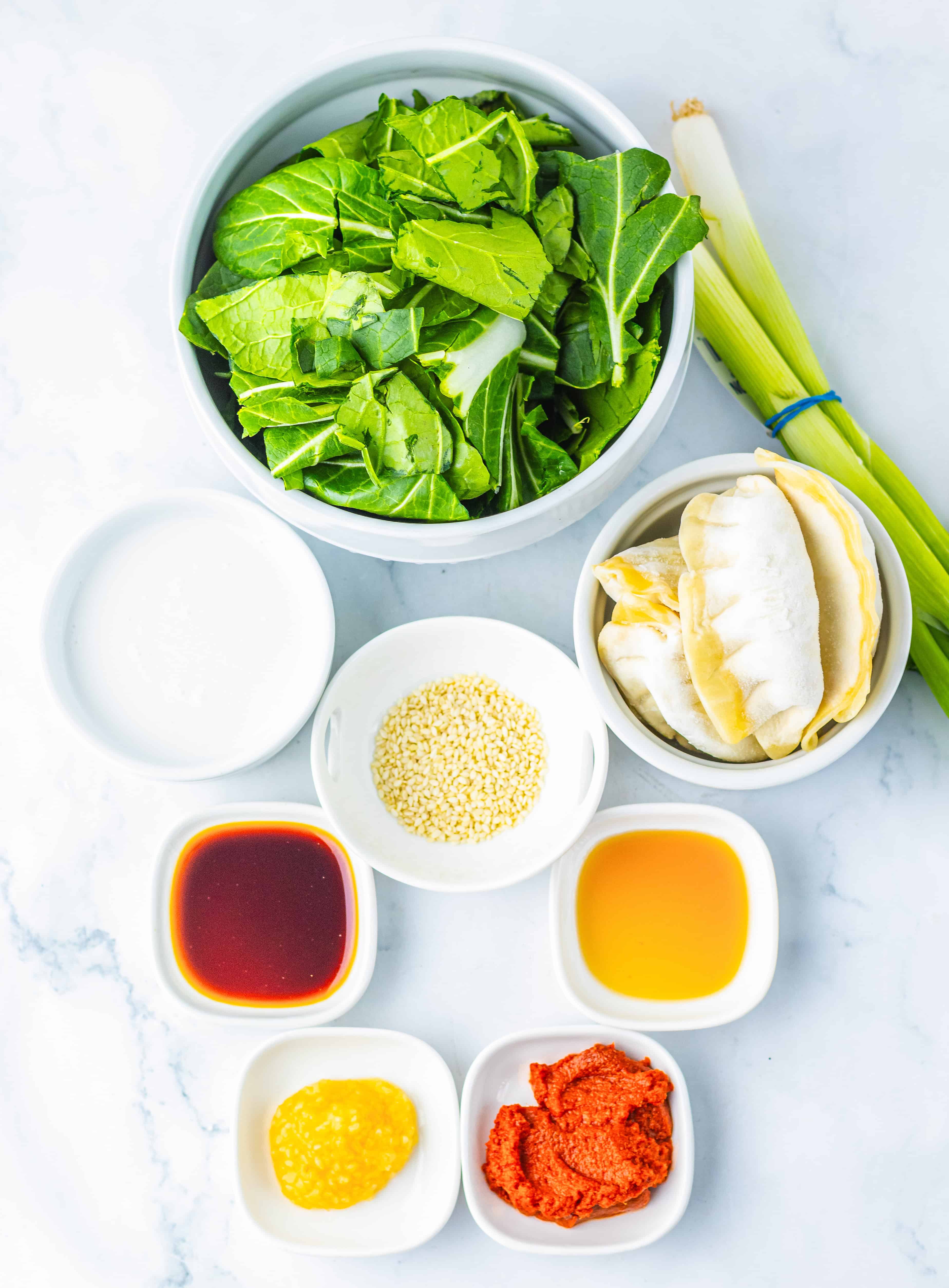 Bowls with greens, sesame seeds, sauces, coconut milk, paste, ginger, scallions, and a tasty Viral Dumpling Bake sit on a white surface.