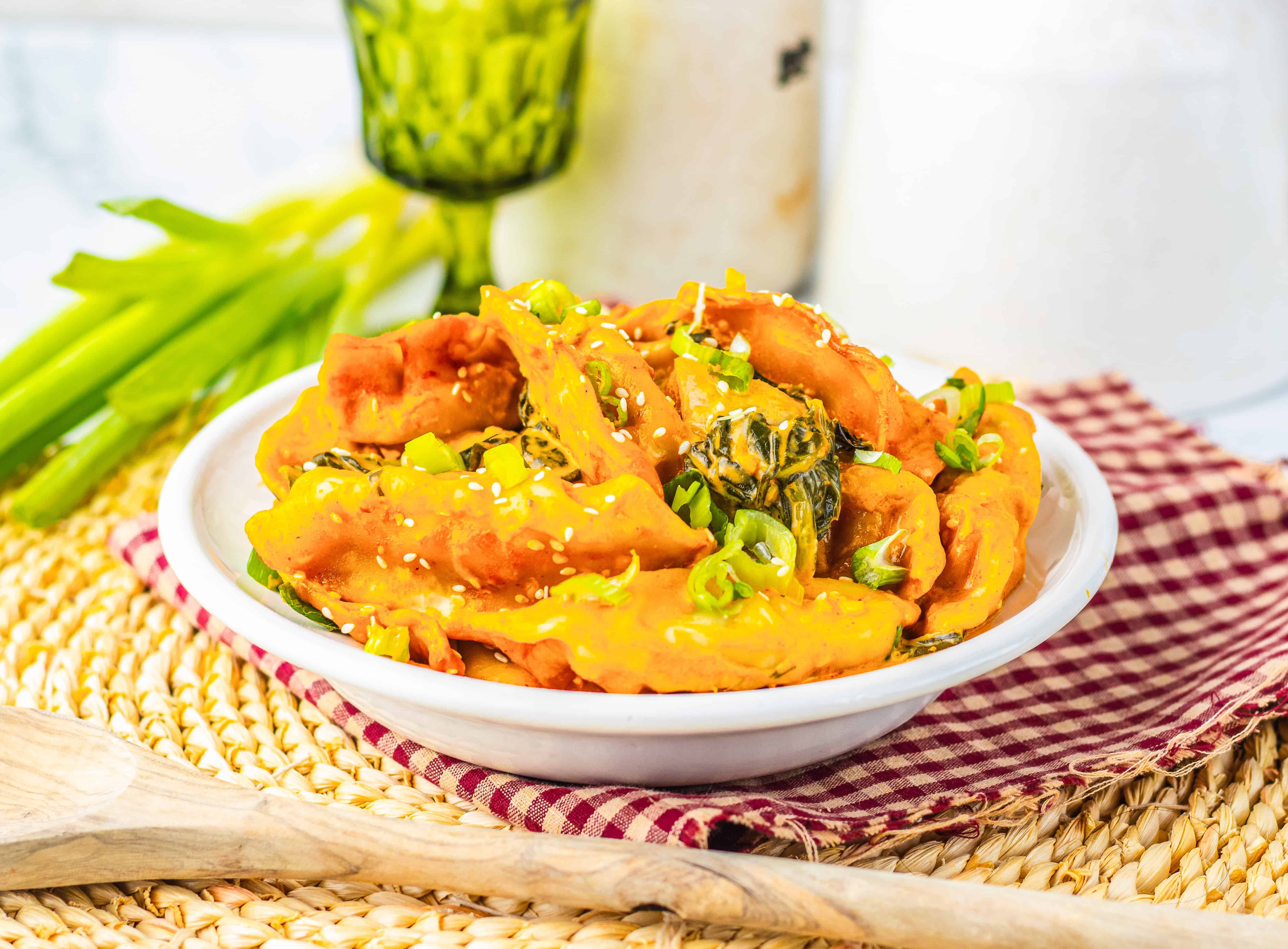 A bowl of spicy dumplings garnished with sesame seeds and green onions sits on a red checkered cloth—perfect for recreating the Viral Dumpling Bake trending on TikTok.