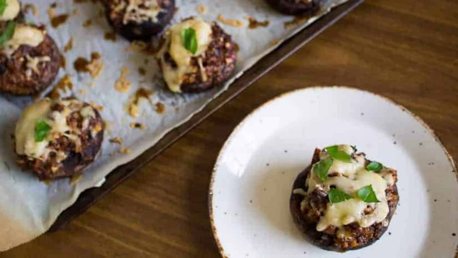 Stuffed mushrooms with melted cheese and herbs on a tray, with one served on a white plate.