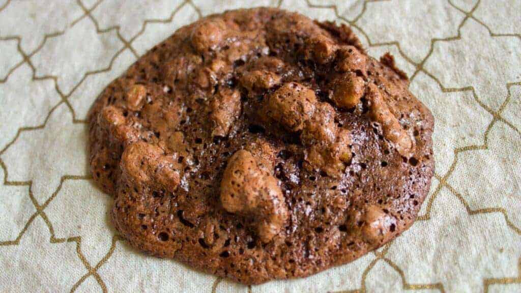A single chocolate walnut cookie sits on a patterned beige surface.