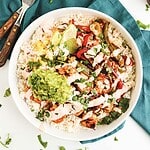 A burrito bowl with rice, grilled chicken, guacamole, corn, peppers, lime, and cilantro on a teal napkin.