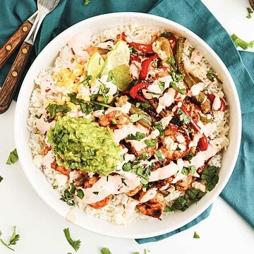 A burrito bowl with rice, grilled chicken, guacamole, corn, peppers, lime, and cilantro on a teal napkin.