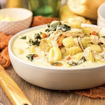 Creamy soup with gnocchi, chicken, spinach, and carrots in a white bowl on a wooden table.