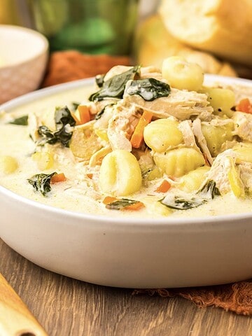 Creamy soup with gnocchi, chicken, spinach, and carrots in a white bowl on a wooden table.