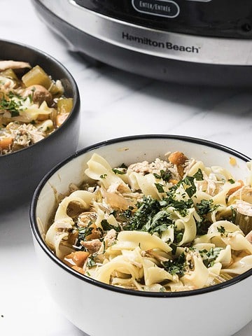 Two bowls of chicken noodle soup garnished with parsley, with a slow cooker in the background.