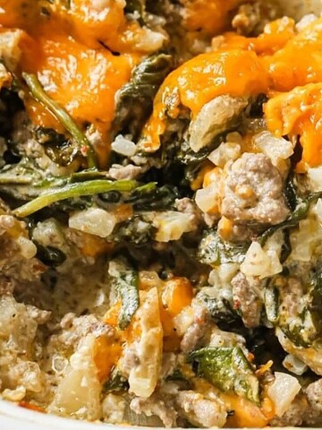 Close-up of a cheesy baked casserole with ground meat, spinach, and onions, with a wooden spoon scooping some.