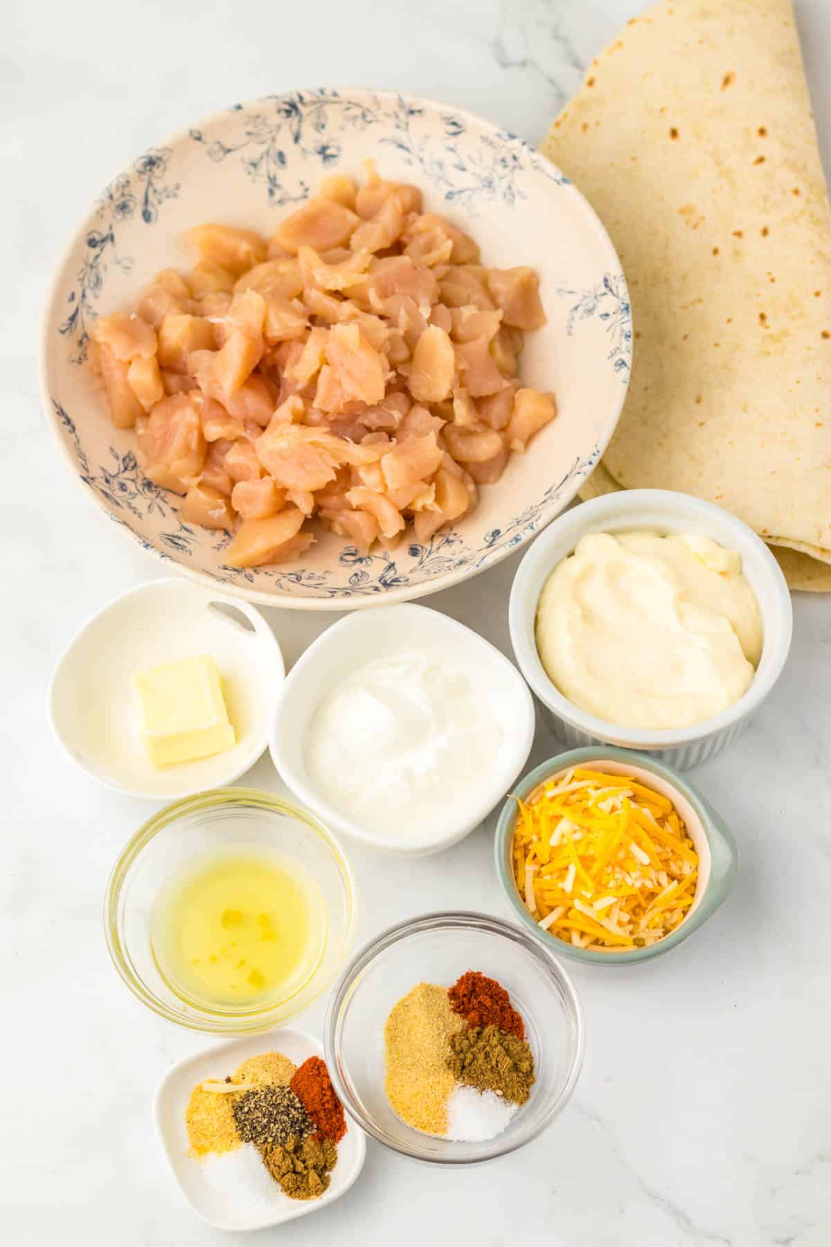 A bowl of diced chicken with tortillas, shredded cheese, butter, sour cream, mayo, oil, and various spices in small bowls—perfect for making a Chicken Quesadilla Copycat just like Taco Bell’s popular favorite.