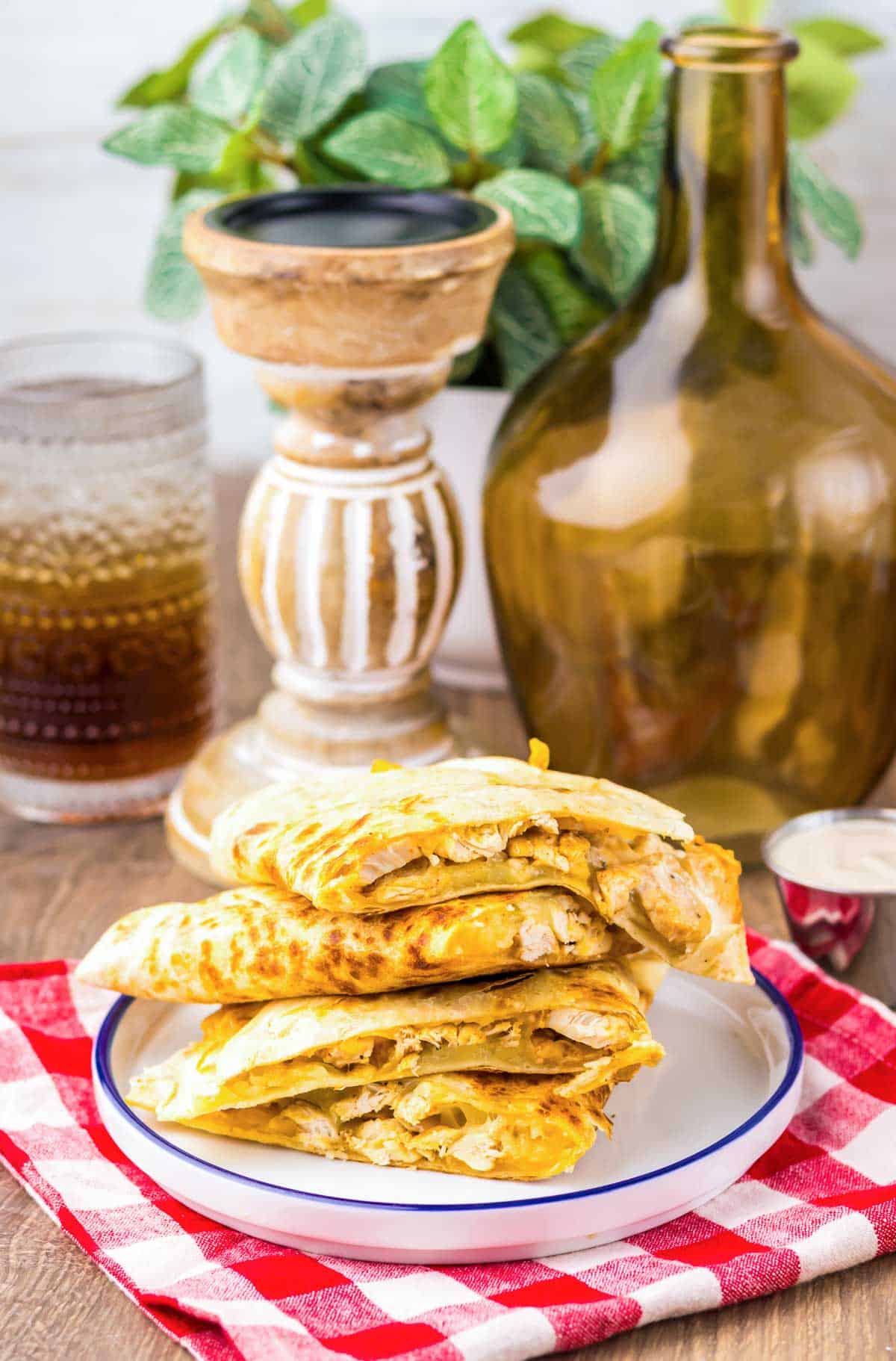 Four Copycat Chicken Quesadillas stacked on a white plate with a drink and decor in the background on a red checkered cloth, just like your favorite Taco Bell treat.