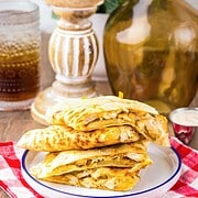 A plate of folded copycat Chicken Quesadillas, inspired by Taco Bell, rests on a red checkered cloth, with drinks and decor in the background.