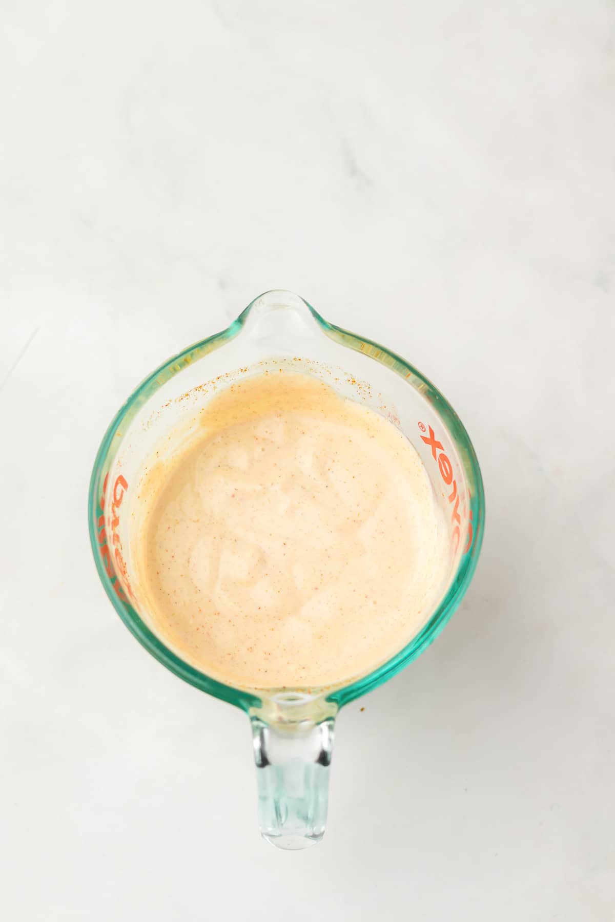 A glass measuring cup with creamy, light orange Copycat Taco Bell Chicken Quesadilla sauce sits on a white surface.