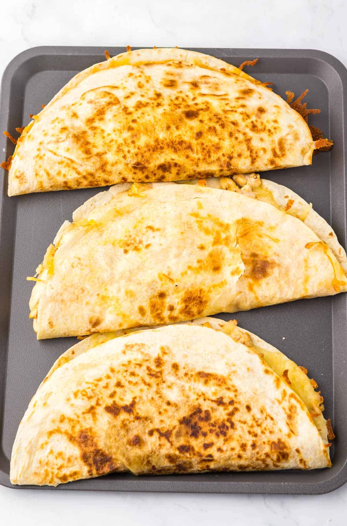 Three golden-brown Copycat Chicken Quesadillas, inspired by Taco Bell, arranged in a row on a dark baking tray.