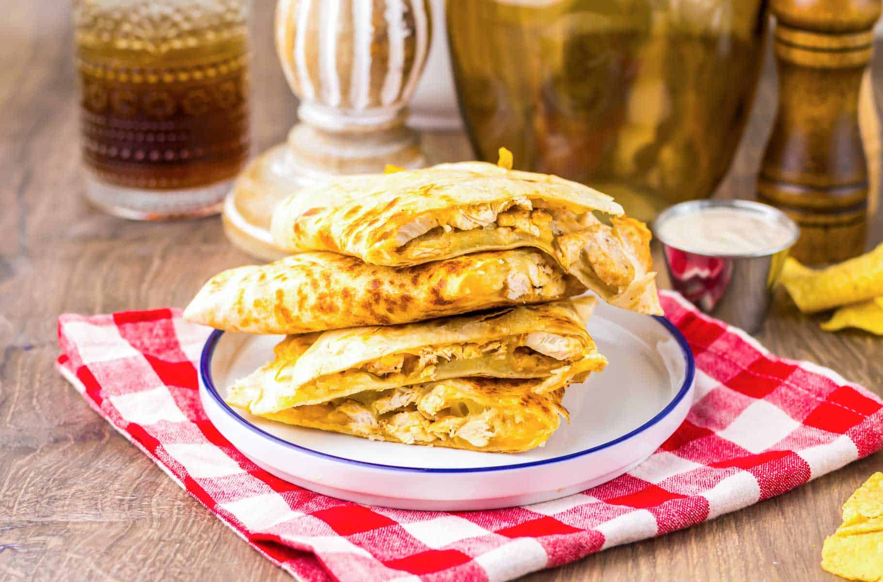 Three copycat Chicken Quesadilla wedges stacked on a plate, served with dipping sauce on a red checkered napkin—just like Taco Bell.