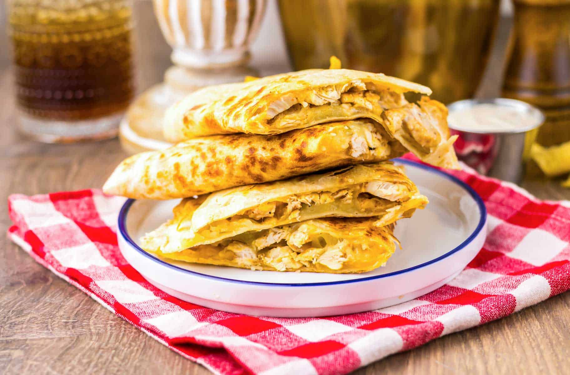 Three copycat Chicken Quesadillas, inspired by Taco Bell, are stacked on a plate and set atop a red and white checkered cloth.