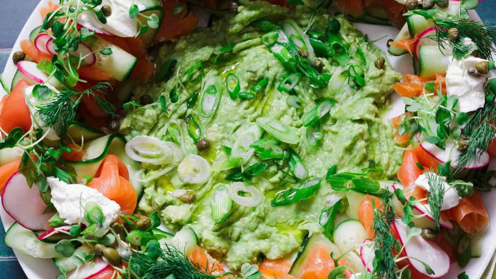 Green avocado dip surrounded by cucumber, radish, smoked salmon, capers, herbs, and cream cheese.