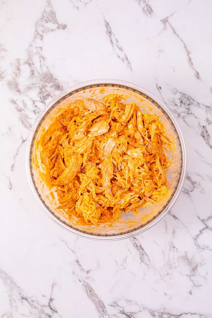 A glass bowl filled with shredded chicken mixed in an orange creamy sauce on a marble surface&mdash;perfect for making Buffalo Chicken Sliders.