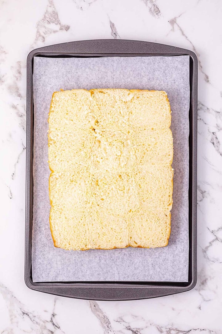 A baking sheet with parchment paper holds sliced bread rolls arranged in a rectangle on a marble surface, perfect for assembling easy Buffalo Chicken Sliders.