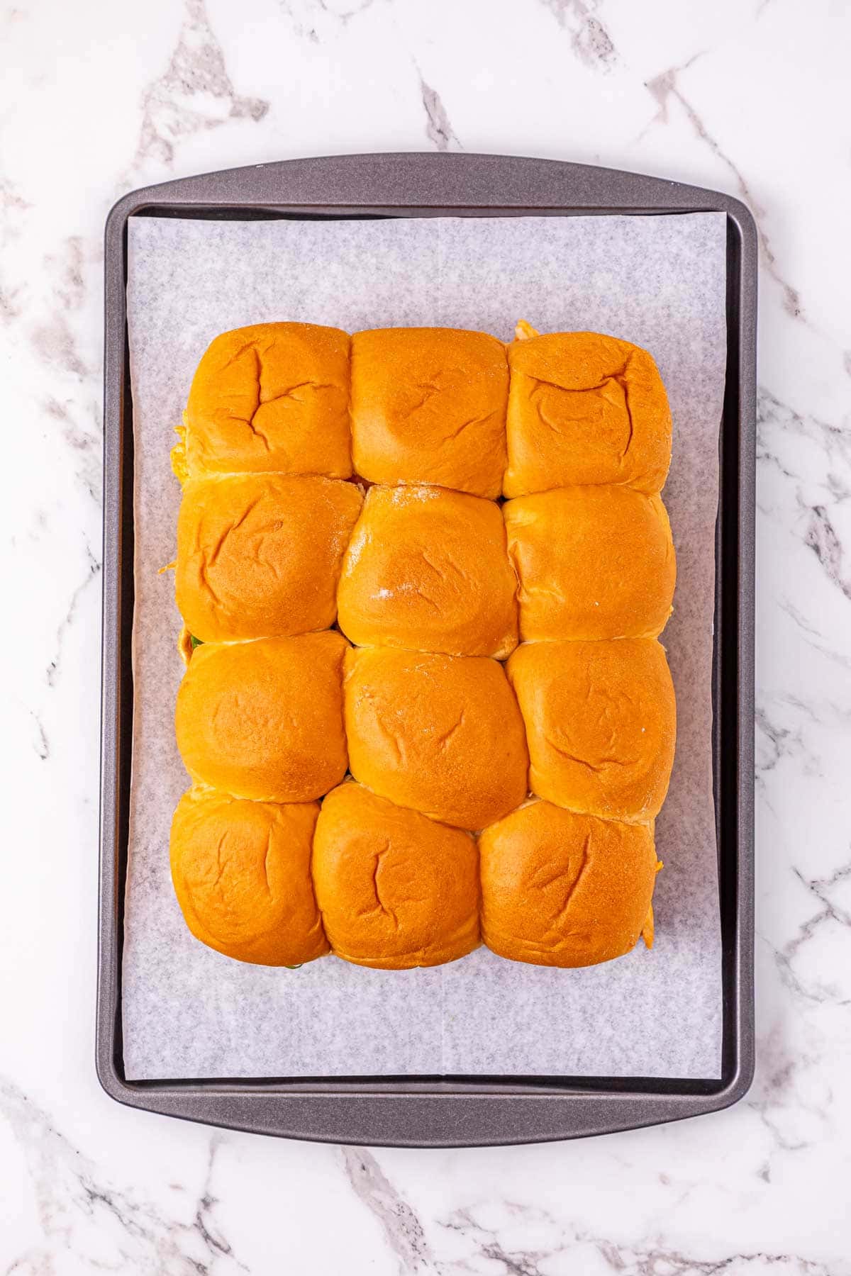 A baking tray lined with parchment paper holds twelve golden brown dinner rolls&mdash;perfect for making Buffalo Chicken Sliders&mdash;on a marble surface.