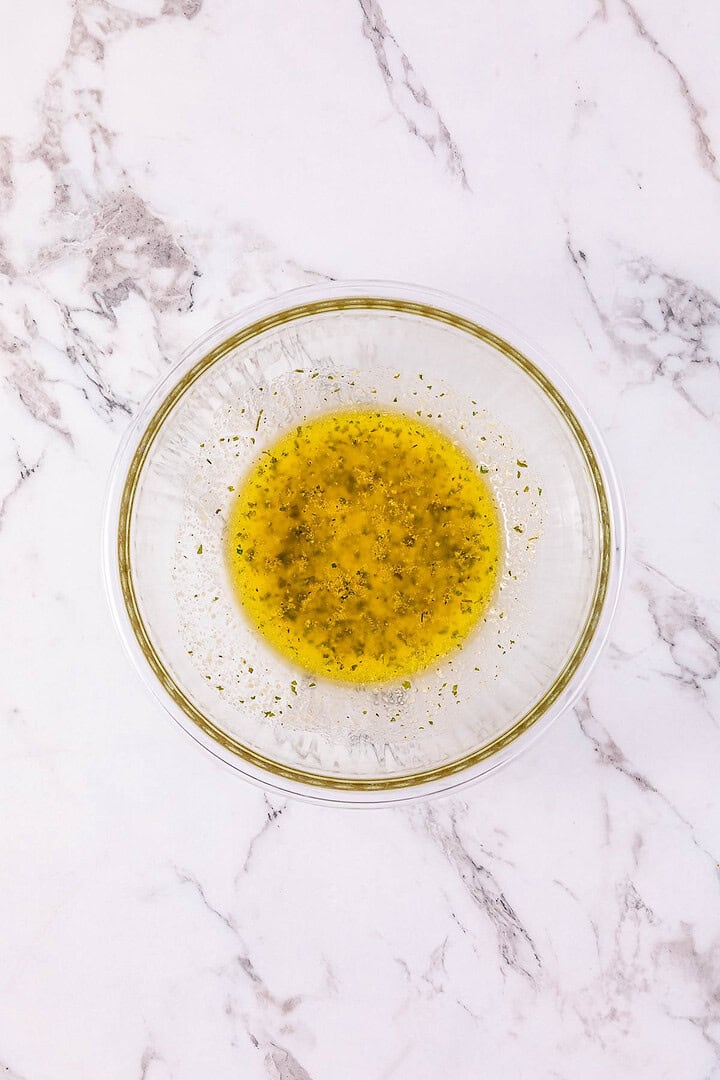 Glass bowl with yellow herb vinaigrette on a white marble surface, perfect for drizzling over Buffalo Chicken Sliders.
