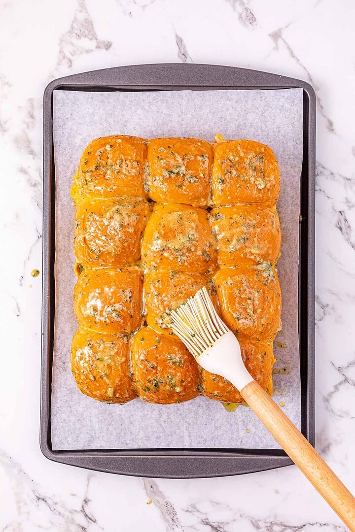 A tray of golden baked rolls brushed with herb butter, perfect for serving as Easy Chicken Sliders, on parchment paper, with a pastry brush on top.
