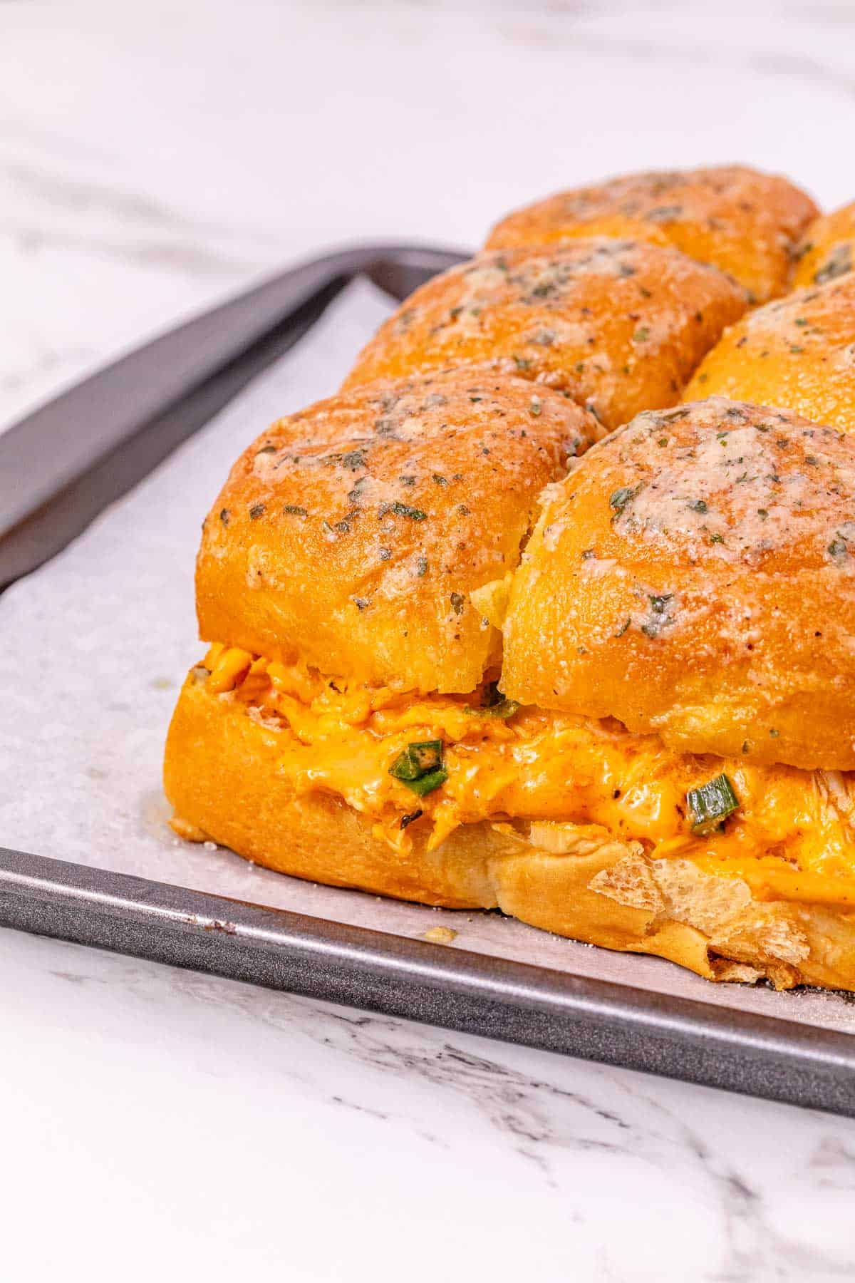 Close-up of cheesy Buffalo Chicken Sliders on golden buns with herbs, resting on a baking tray lined with parchment paper.