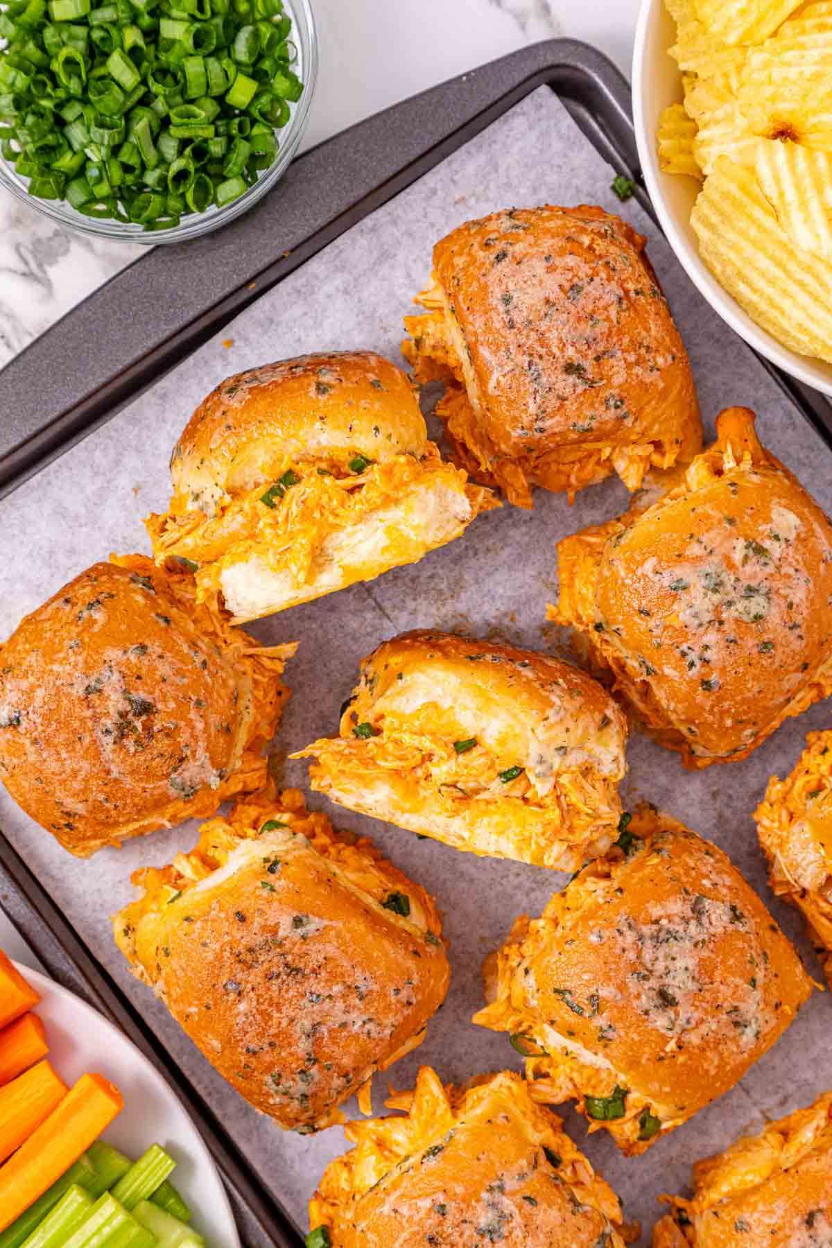 Buffalo Chicken Sliders with shredded chicken and cheese on a tray, surrounded by chips, green onions, and vegetables&mdash;these easy sliders are perfect for any gathering.