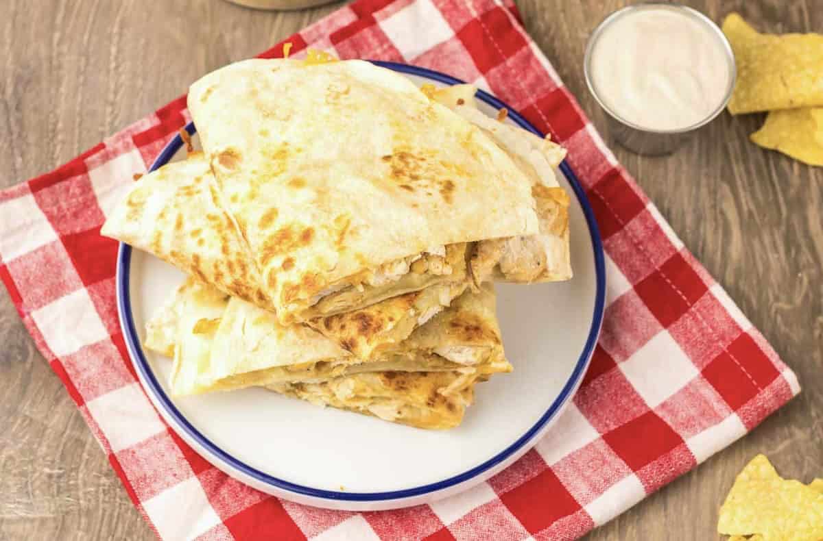 Stack of cheesy Chicken Quesadilla wedges on a white plate, with dip on the side, served atop a red checkered cloth—your own Taco Bell copycat creation at home.