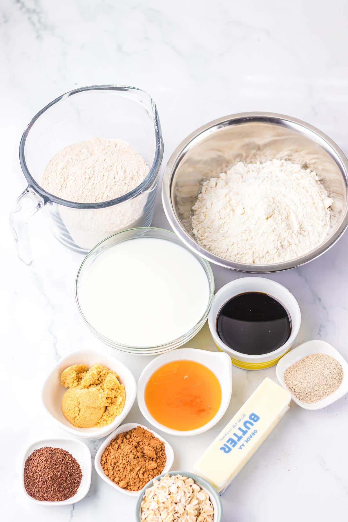 Various baking ingredients in bowls, including flour, milk, butter, oats, and molasses on a white surface—perfect for recreating the famous Cheesecake Factory Brown Bread recipe at home.