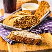 Slices of Cheesecake Factory Brown Bread, one with butter, are arranged on a wooden board, accompanied by a drink and sauce in the background. Try this delicious recipe at home!.
