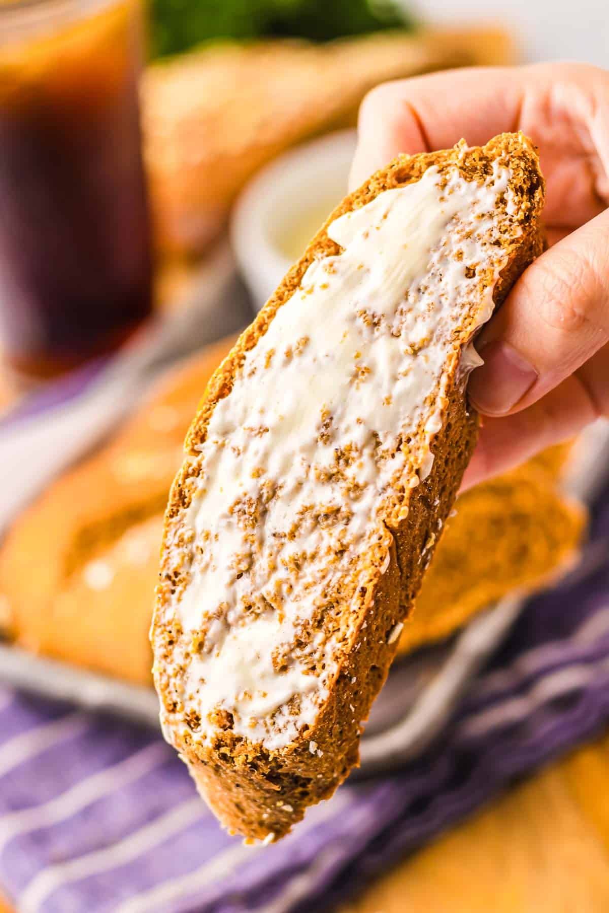 A hand holds a slice of Cheesecake Factory brown bread spread with butter, with more bread and a drink in the background.