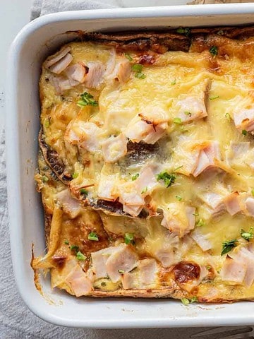 Baked casserole topped with melted cheese, chopped ham, and fresh herbs in a white dish on a woven mat.
